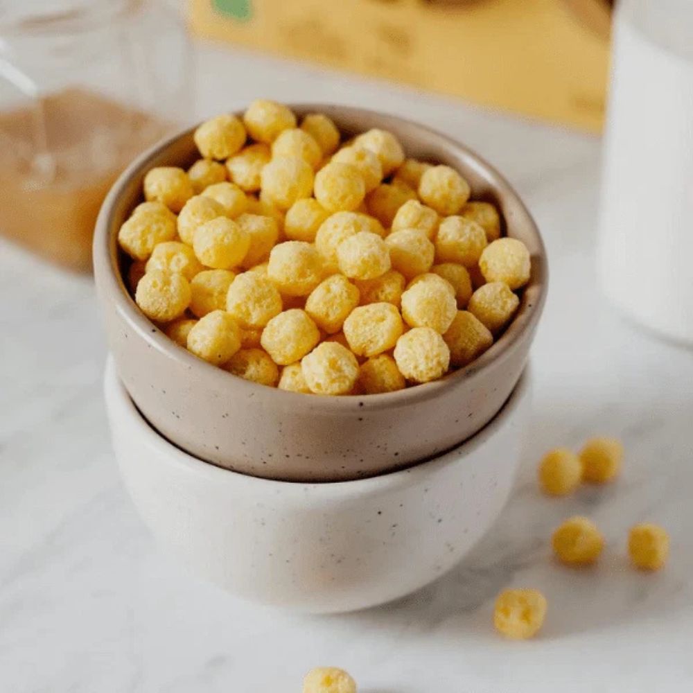 A stack of two speckled ceramic bowls filled with small yellow cereal puffs, set on a light marble surface with a few puffs scattered nearby.