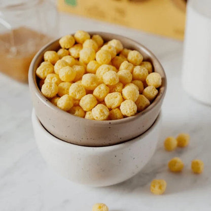 A stack of two speckled ceramic bowls filled with small yellow cereal puffs, set on a light marble surface with a few puffs scattered nearby.