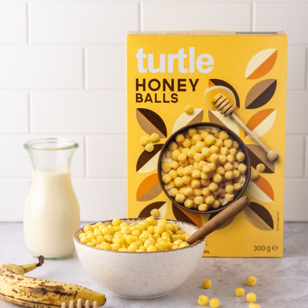 A box of Turtle Honey Balls cereal stands behind a bowl filled with yellow cereal balls, with a glass of milk and a banana on a kitchen counter against a white tiled wall.