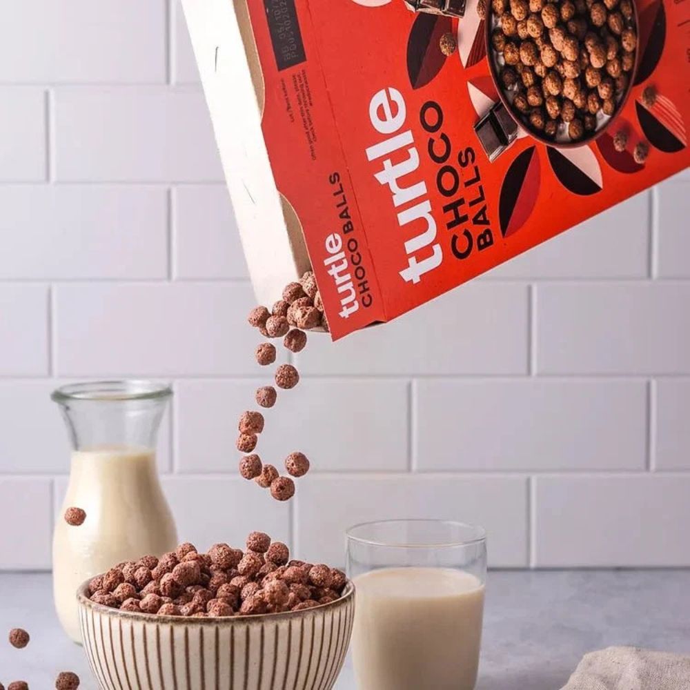 A red box of turtle CHOCO BALLS cereal is tilted over a striped bowl, with cereal balls pouring in midair. A glass and a glass carafe of milk are placed on a countertop against a white subway tile backsplash.