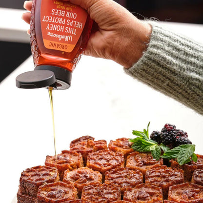 Hand pouring honey from a bottle onto a dessert with berries and mint leaves.