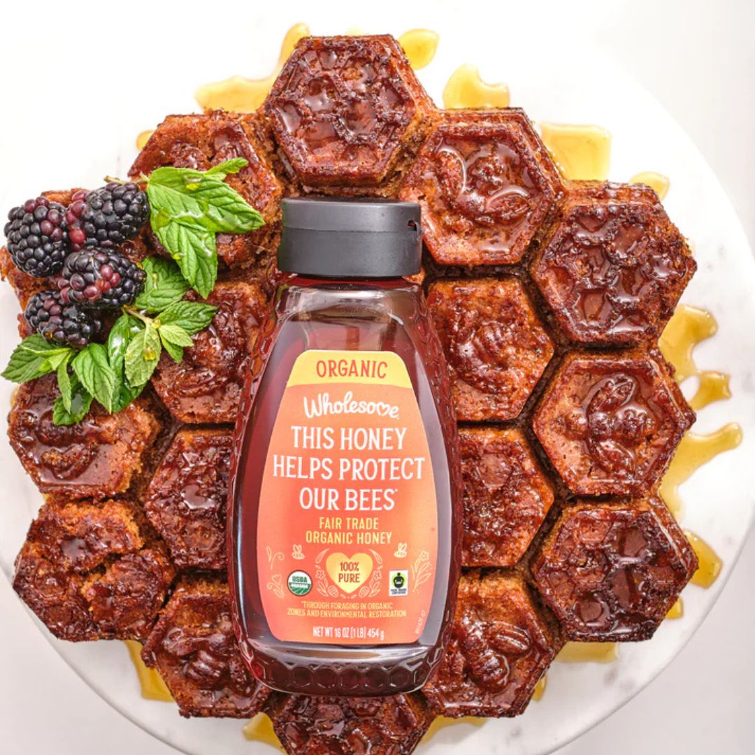 Honeycomb with a bottle of Wholesome Sweeteners honey surrounded by berries and mint leaves on a white background.