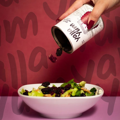 Hand pouring dried berries from a can labeled 'Willa' onto a salad bowl against a red background with blurred text.