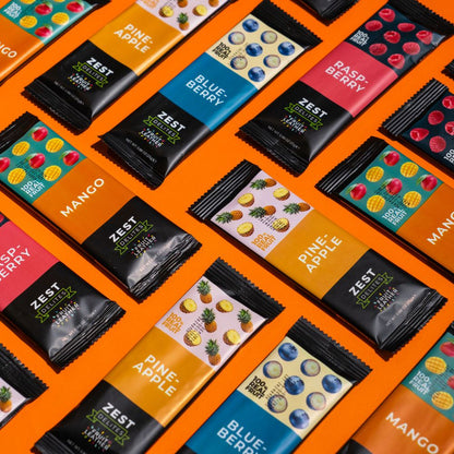 Multiple Zest Nutrex protein bars in individual wrappers arranged diagonally on a bright orange background. Visible flavors include Mango, Pine-Apple, Blue Berry, Raspberry, and Strawberry, with colorful fruit illustrations and black-and-colored packaging.